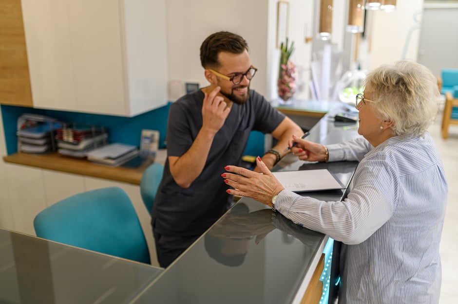 Optometry front-desk staff assisting an older female patient with paperwork at the check-in counter.
